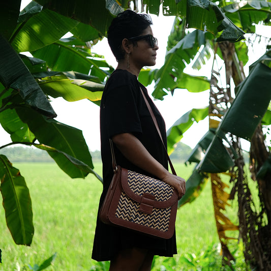 A person standing in a natural setting with green foliage in the background, wearing a black dress and carrying a brown and blue zigzag patterned sling bag.